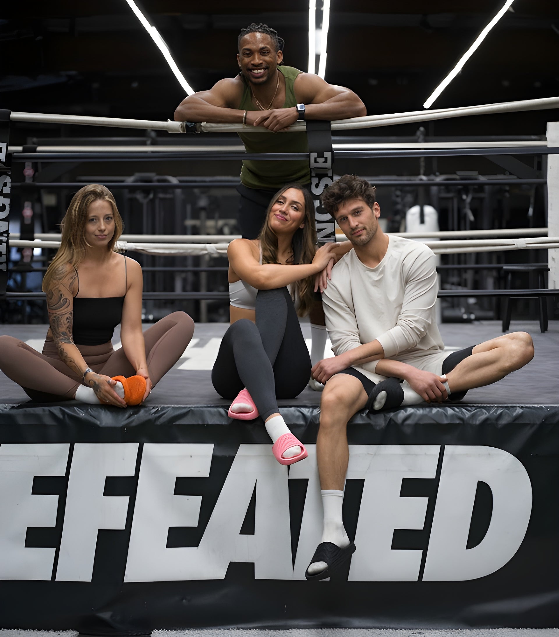 Four people sitting on a boxing ring with a 'DEFEATED' banner